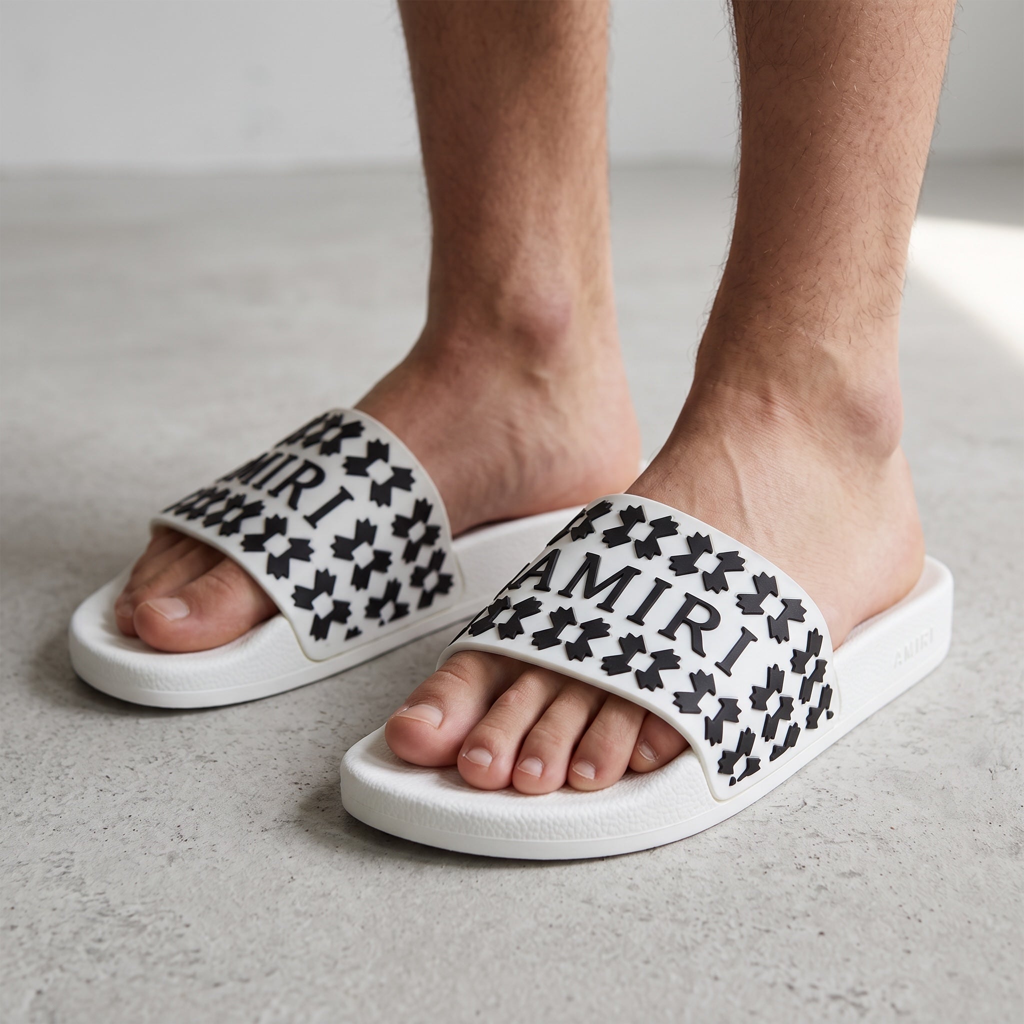 Close-up of white Amiri logo slide sandals with black raised logo pattern on model's feet.