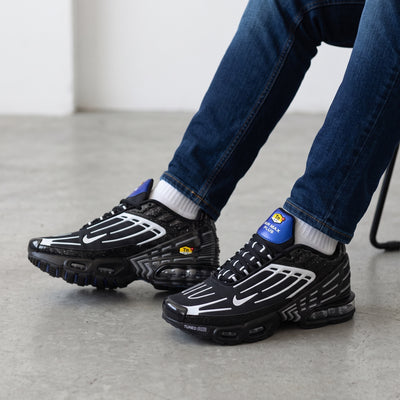 Pair of black Nike Air Max Plus Tuned Air sneakers with white overlays and blue heel tab