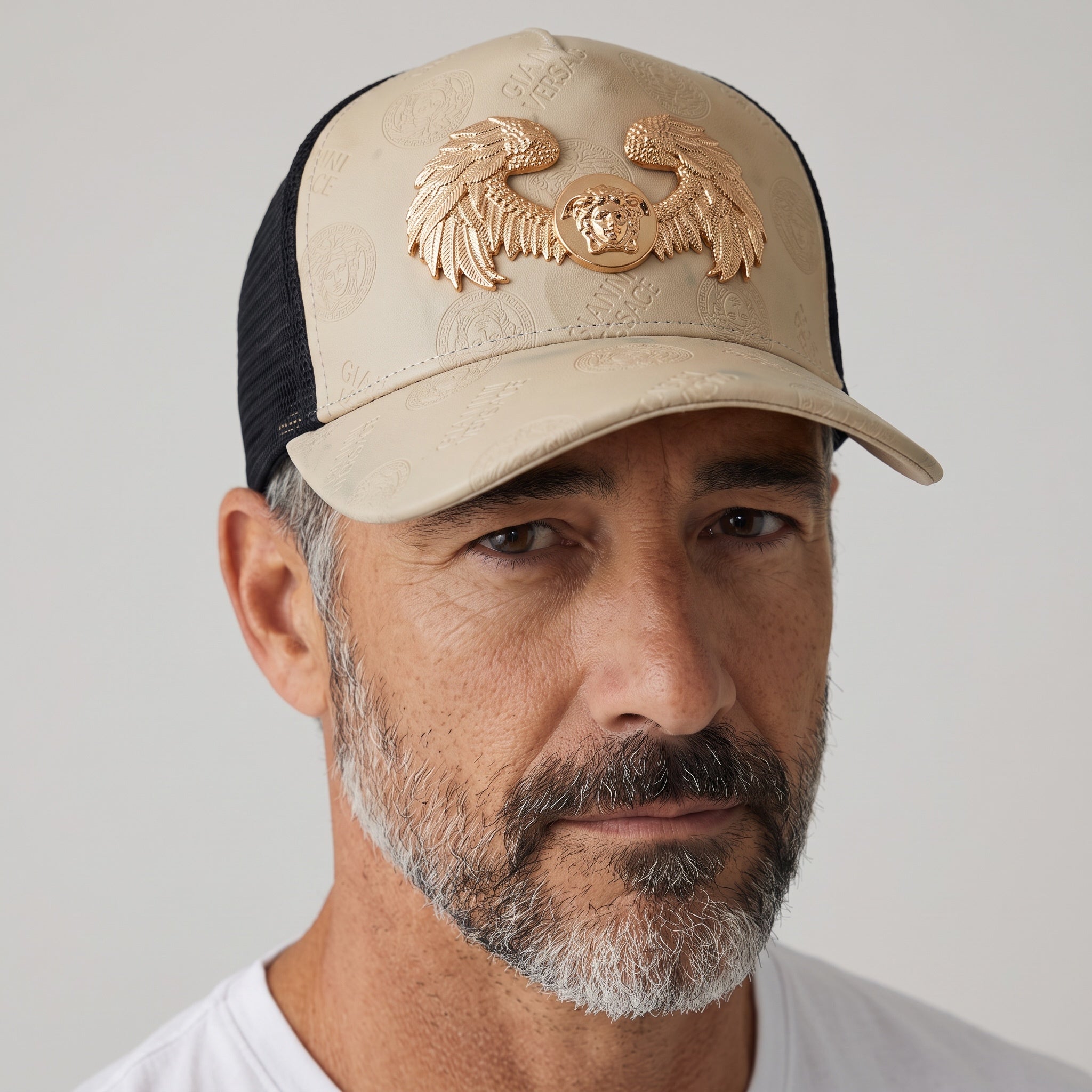 Man wearing a beige and black trucker cap with an embossed logo pattern and a gold Medusa wings emblem on the front