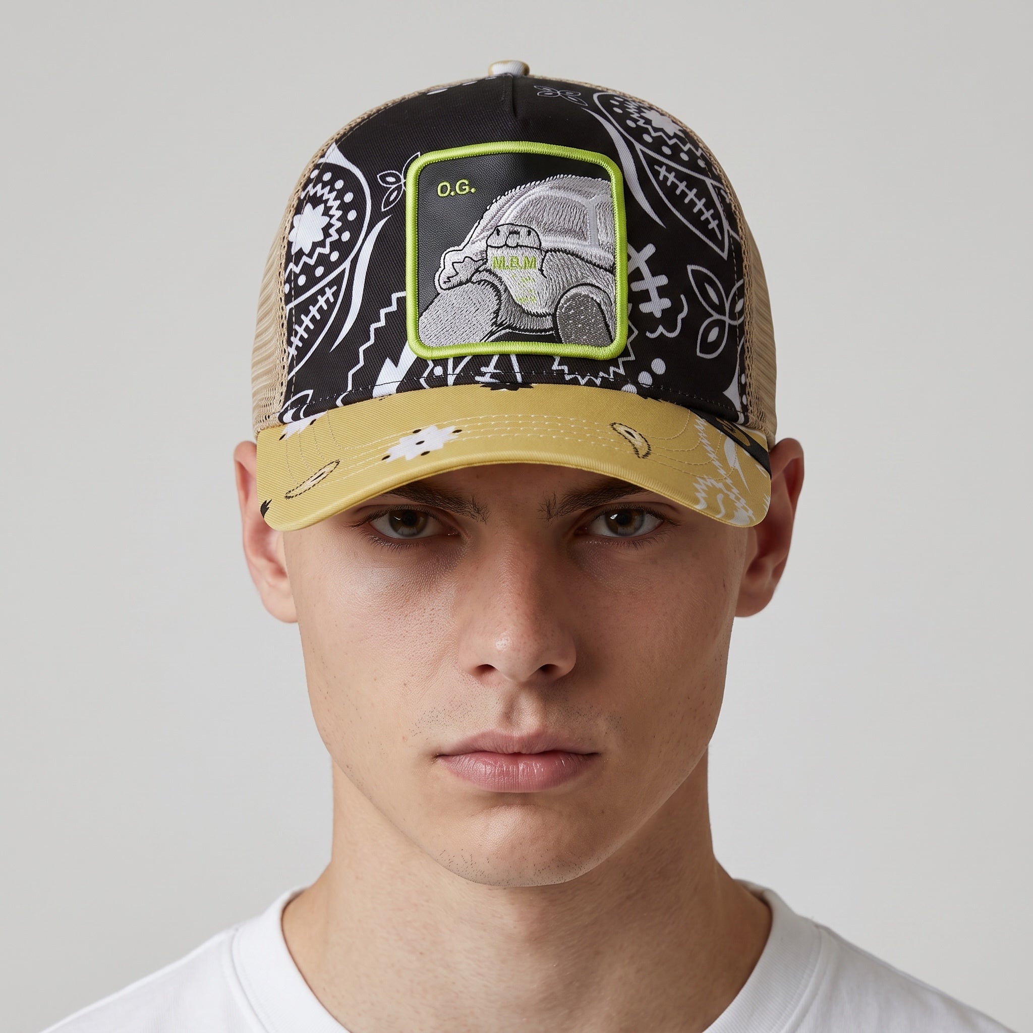 Front view of a trucker cap with black bandana print, embroidered square patch reading O.G. and a yellow brim