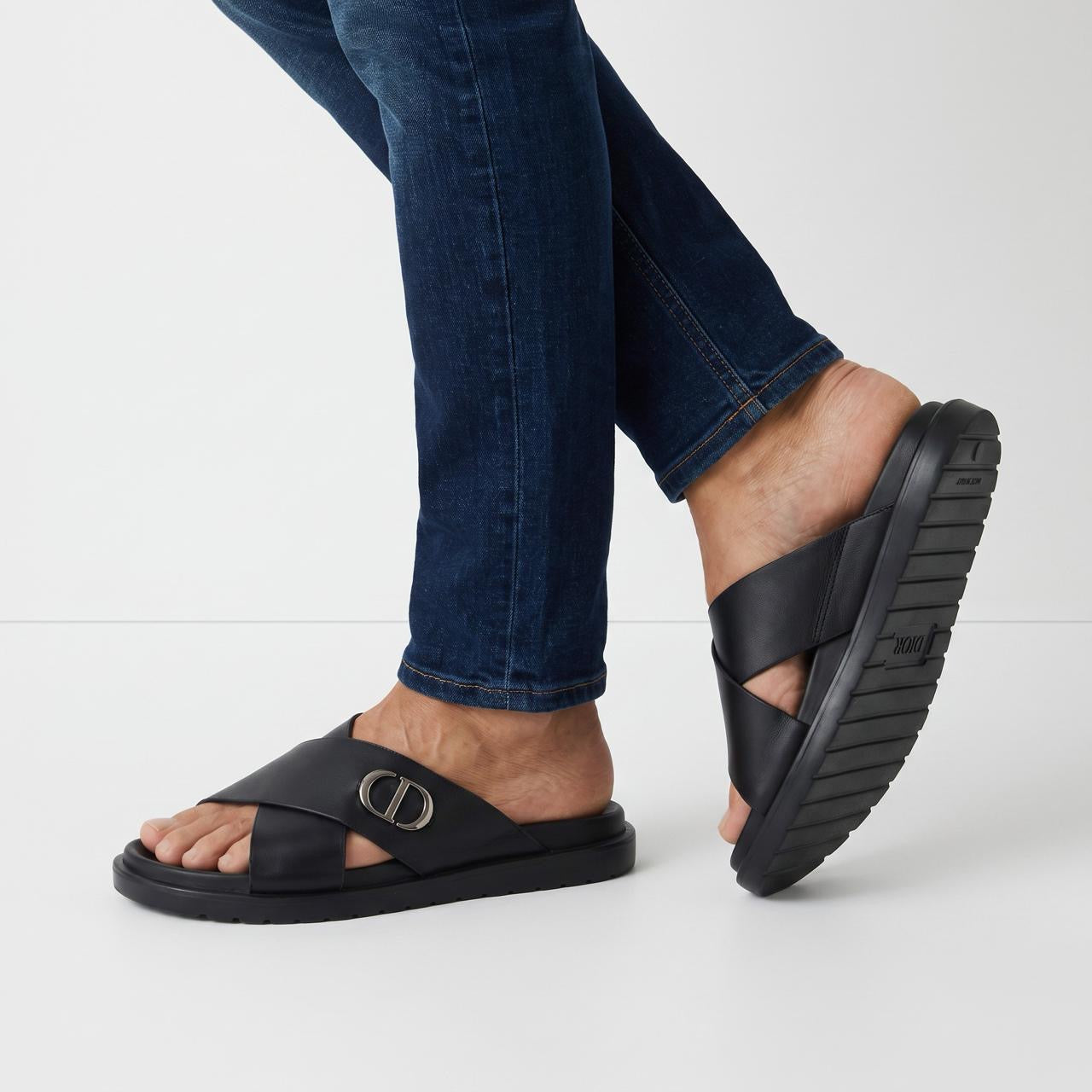 Black Dior crisscross slide sandal with metal CD logo and textured rubber sole, shown on model feet