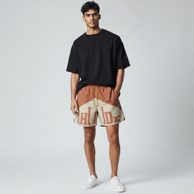 MASTERPIECE Rhude panel swim shorts in rust brown and cream with oversized logo detail and yellow drawstring. Model wears black t-shirt and white sneakers.